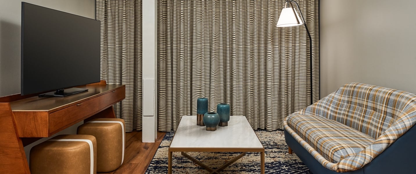 Modern living area featuring a large TV, a coffee table with three blue vases, and a plaid sofa. Wavy-striped curtains cover the back wall.