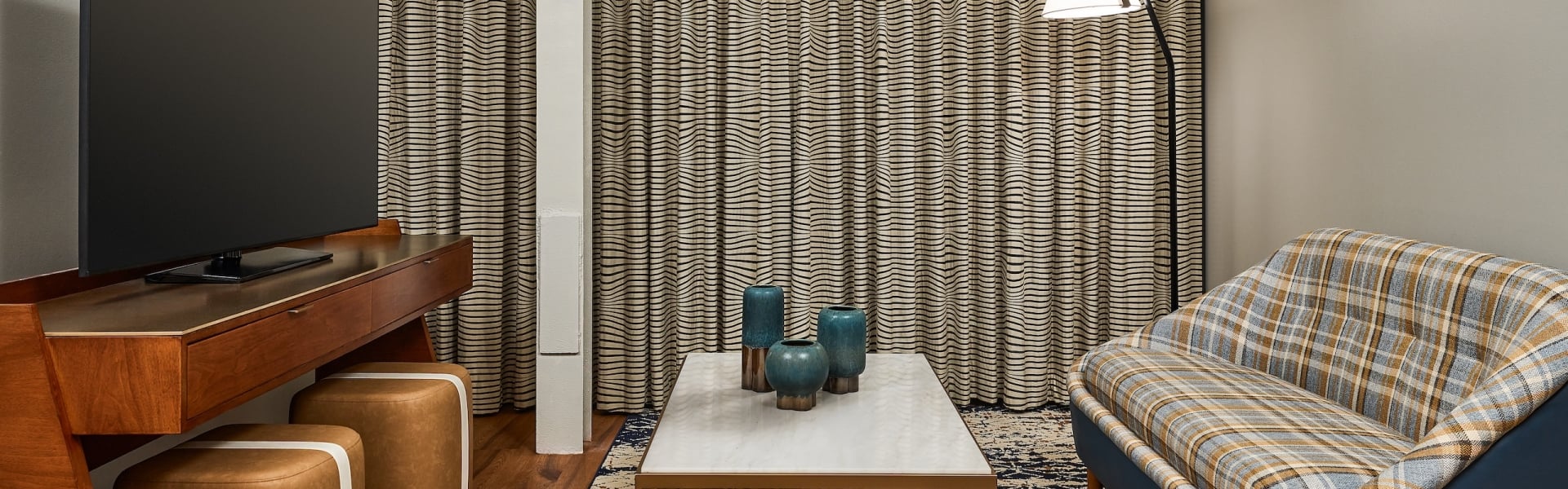 Modern living area featuring a large TV, a coffee table with three blue vases, and a plaid sofa. Wavy-striped curtains cover the back wall.