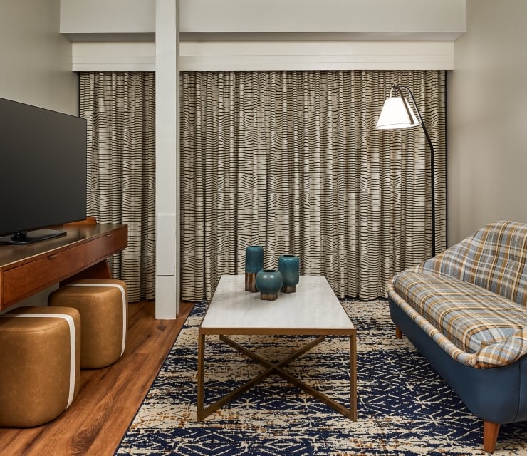 Modern living area featuring a large TV, a coffee table with three blue vases, and a plaid sofa. Wavy-striped curtains cover the back wall.