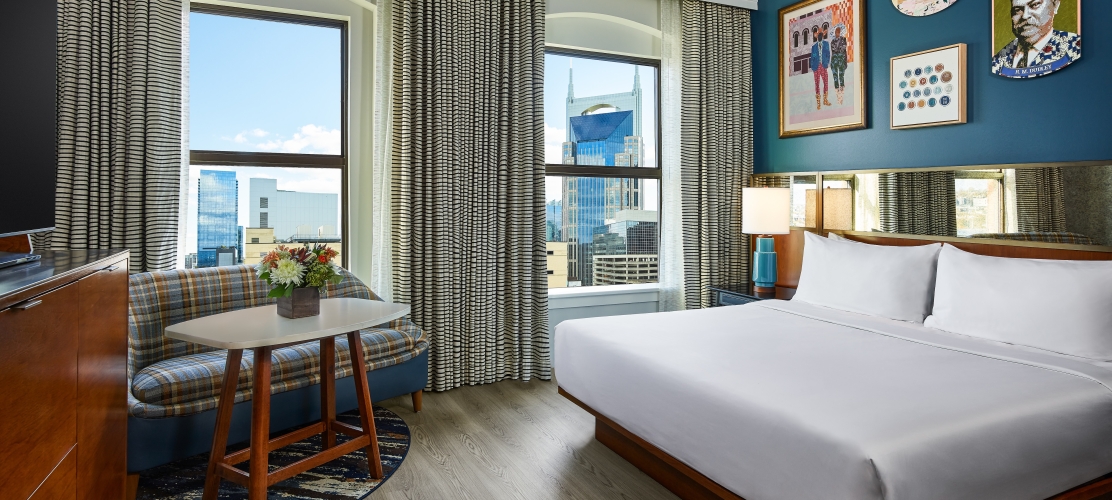 An Untitled Nashville Suite, featuring a King bed against an accent wall with artwork, a loveseat, a large TV and two bright windows overlooking the Nashville skyline
