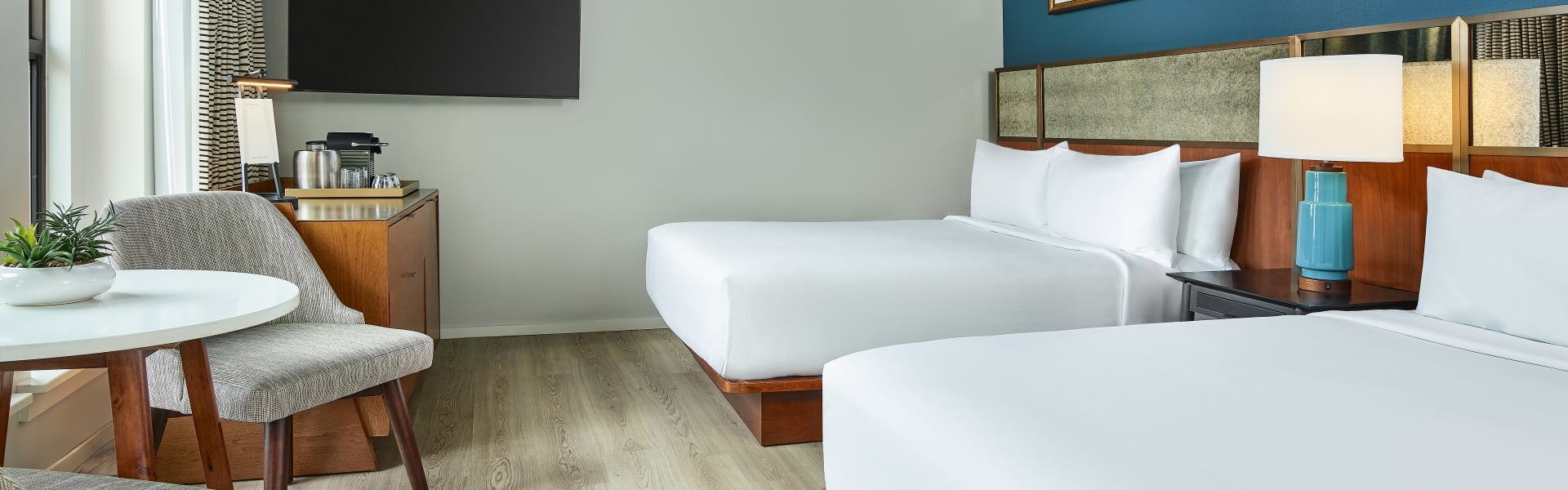 A guestroom at the Untitled Nashville with two queen beds facing a bright window, with a seating area, accent wall featuring artwork and large wall-mounted TV