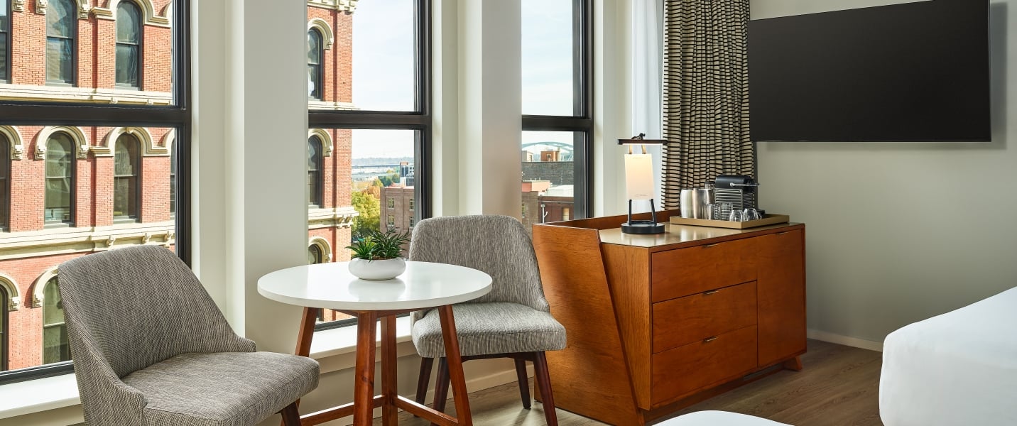 A guestroom at the Untitled Nashville with two queen beds facing a bright window looking out on the Nasvhille skyline, with a seating area and large wall-mounted TV