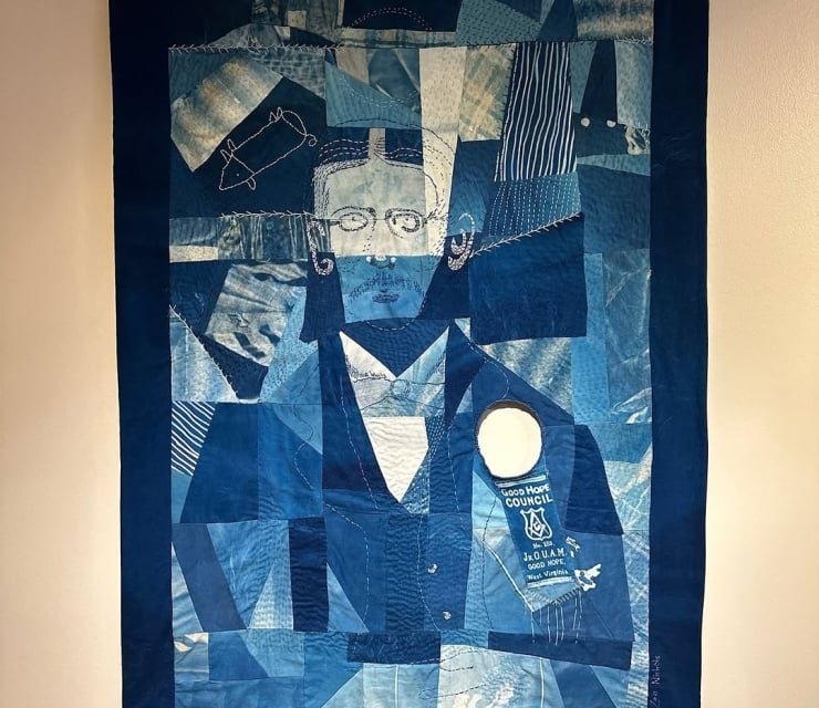 A blue denim patchwork quilt art piece hanging on a wall, featuring a stitched portrait of a person with glasses and a circular cutout showing a vintage ribbon badge.