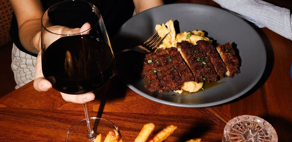 A diner in a black top holding a glass of red wine at a wooden restaurant table featuring sliced steak on mashed potatoes, a metal cup of french fries, and a burger.