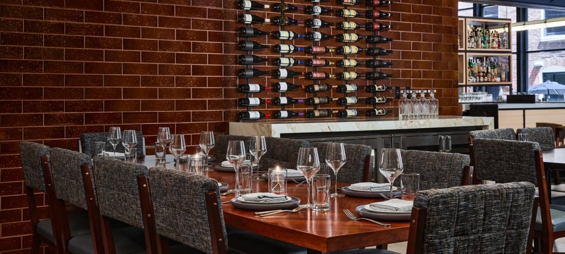 A long wooden dining table set for a group in a restaurant, featuring textured grey chairs and a large wall-mounted wine rack against a dark brick background.