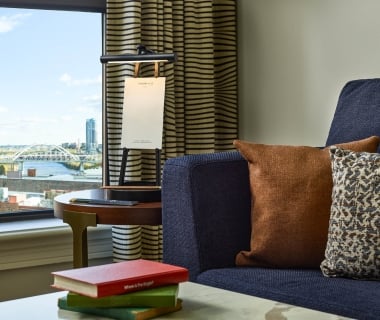 A cozy hotel guest room seating area featuring a blue sofa with throw pillows next to a window overlooking the Cumberland River and the John Seigenthaler Pedestrian Bridge.