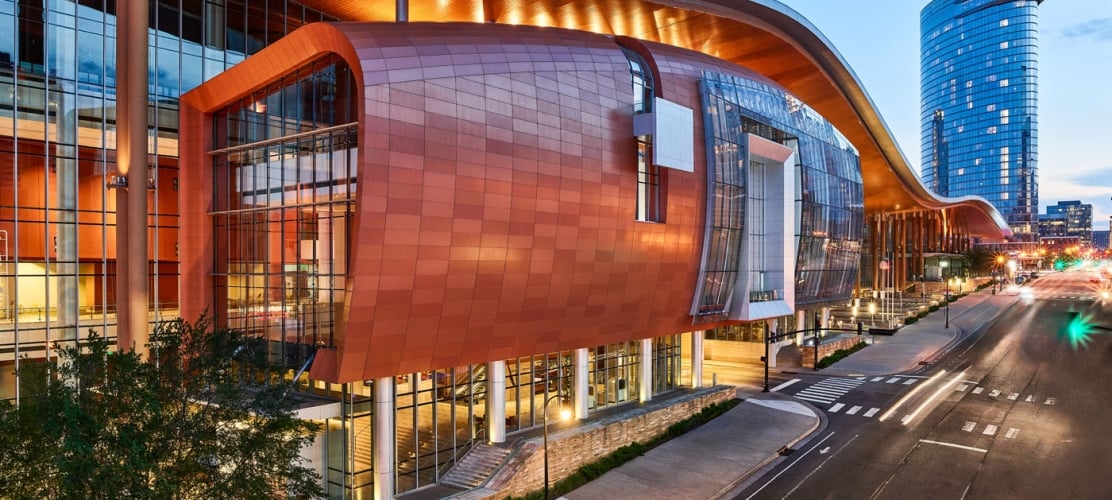 An architectural dusk shot of the modern Music City Center building in downtown Nashville, featuring a sweeping copper-colored exterior and illuminated glass facades along a city street.