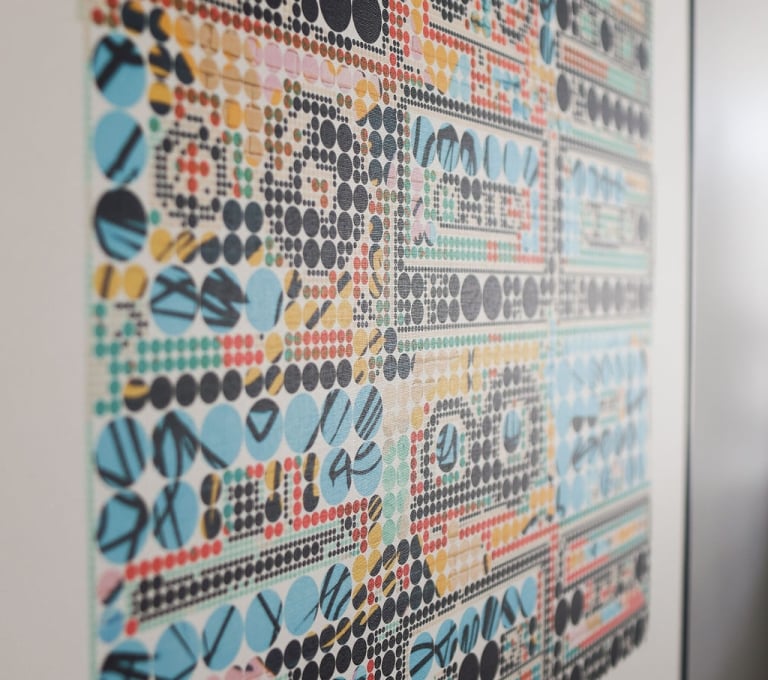 An angled close-up of a framed graphic art print on a wall, featuring vibrant geometric patterns and dot matrices arranged in rows to form abstract shapes.