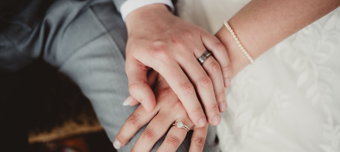 Close-up of a bride and groom holding hands, showcasing a diamond engagement ring, a textured wedding band, a pearl bracelet, and a grey suit.
