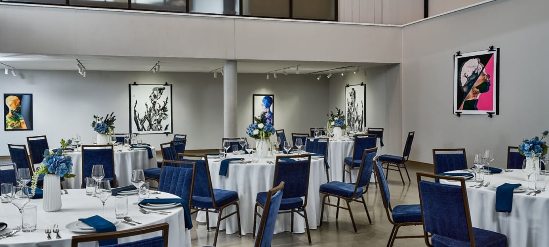 Round dining tables are formally set with white linens, blue chairs, and floral centerpieces, within a spacious, modern gallery-style room featuring various abstract artworks on light gray walls.