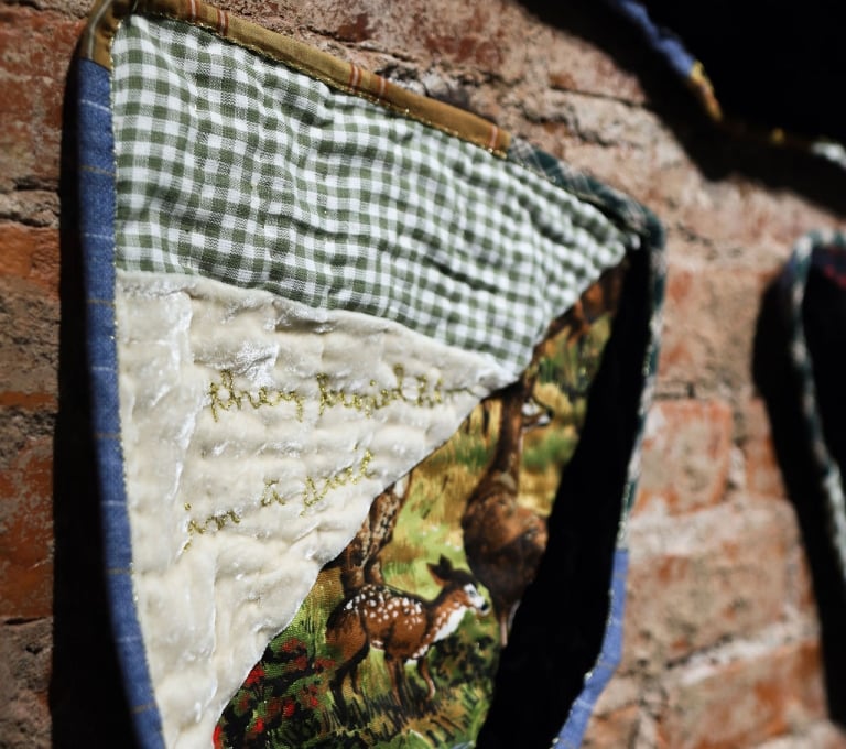 A close-up, angled shot of a handmade textile art piece with green gingham and quilted panels featuring a painted deer, mounted on an exposed red brick wall.