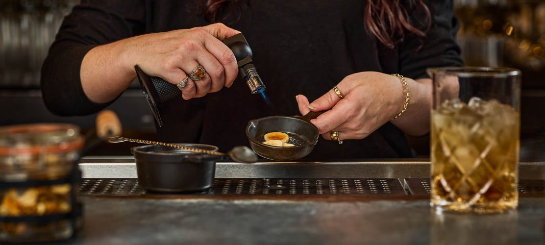 A bartender using a handheld torch to caramelize a garnish in a small dish next to a cocktail glass filled with ice on a bar counter.