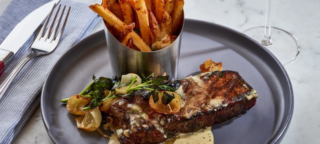 A grilled steak topped with melted herb butter, served with a side of thick-cut french fries in a metal cup and roasted onions on a grey plate.