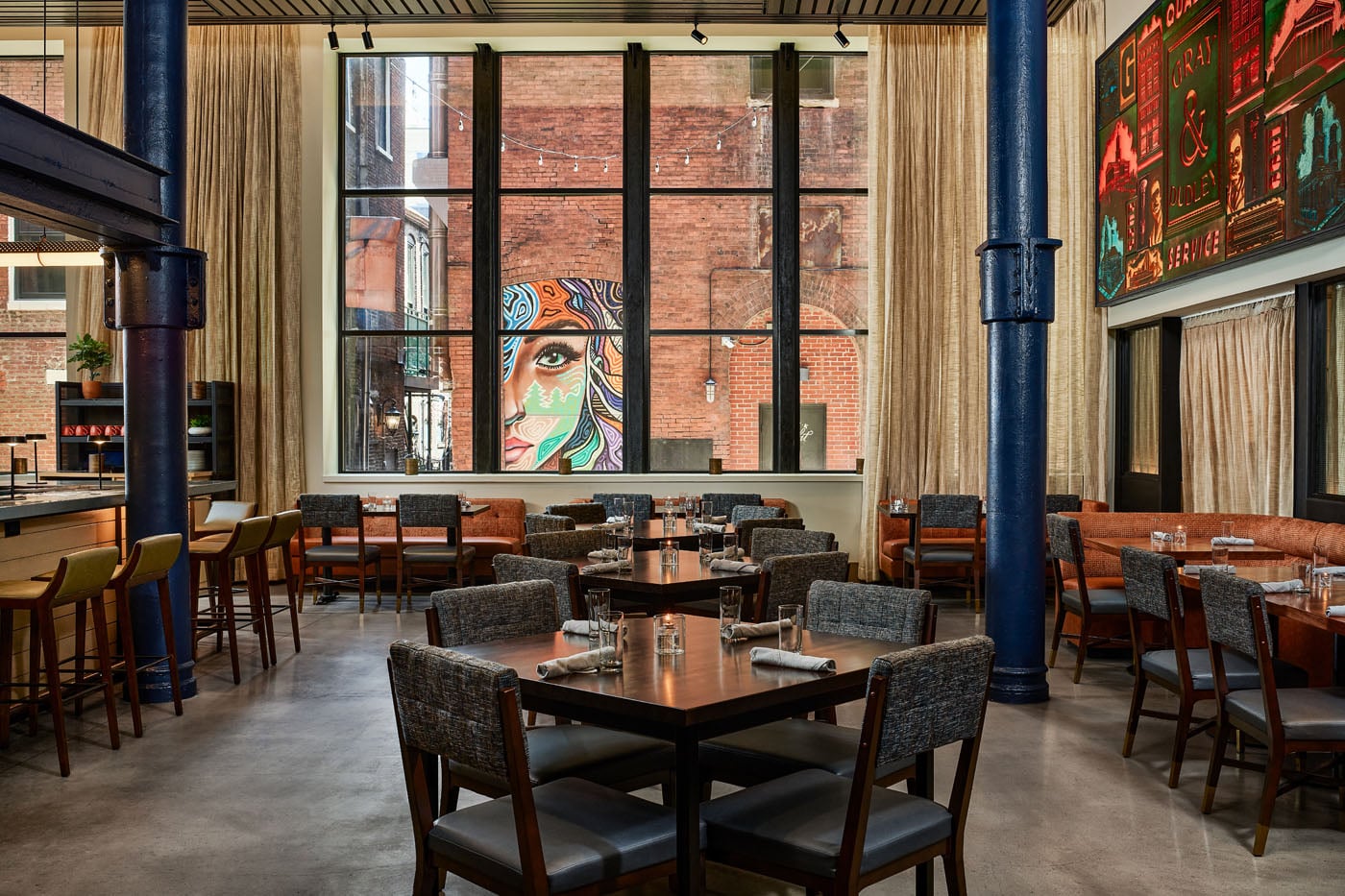 A modern, spacious restaurant dining room featuring dark wood tables, grey textured chairs, tall industrial windows, and striking dark blue structural pillars.