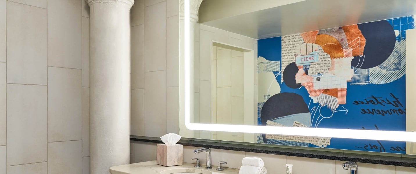 An illuminated mirror reflects a bathroom vanity with a sink, tissue box, and towels. It also shows a blue wall with abstract collage art, including reversed text "TREAE" and "sintaxis armonia".