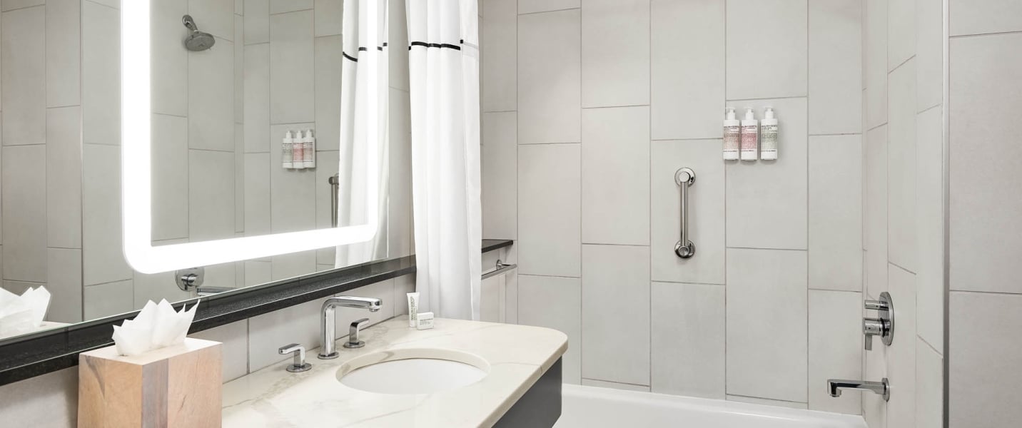 Modern bathroom with marble vanity, lighted mirror, and shower/tub. Wall-mounted dispensers labeled "SHAMPOO", "CONDITIONER", "BODY WASH" are in the shower.