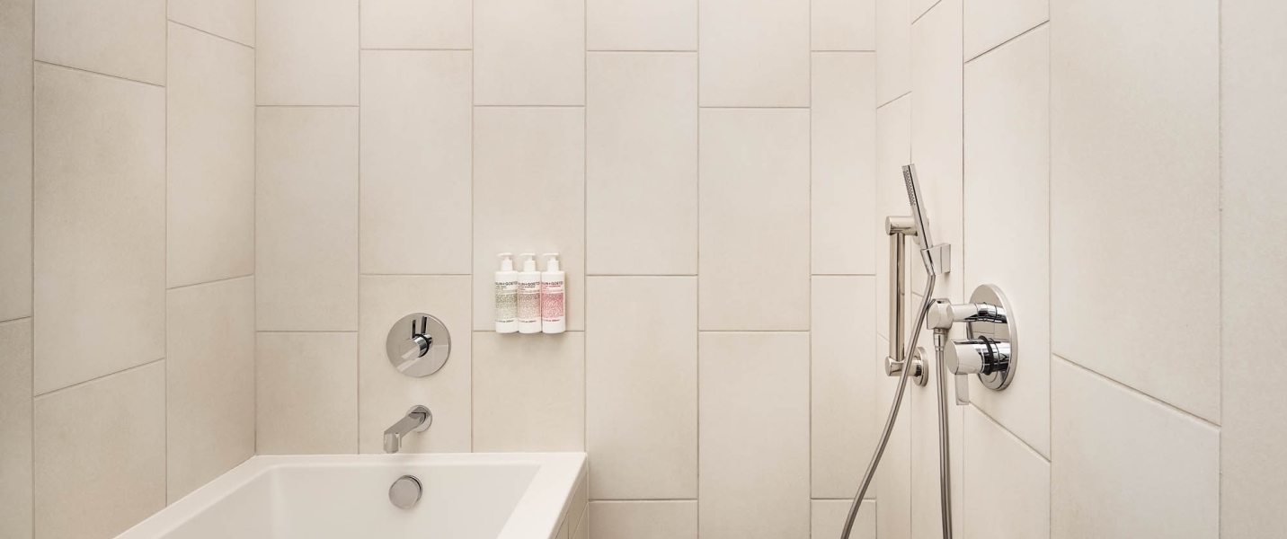A sleek, light-colored tiled bathroom features a white bathtub, chrome fixtures, and three wall-mounted "SUN + GOTO" pump bottles: green, white, and pink.
