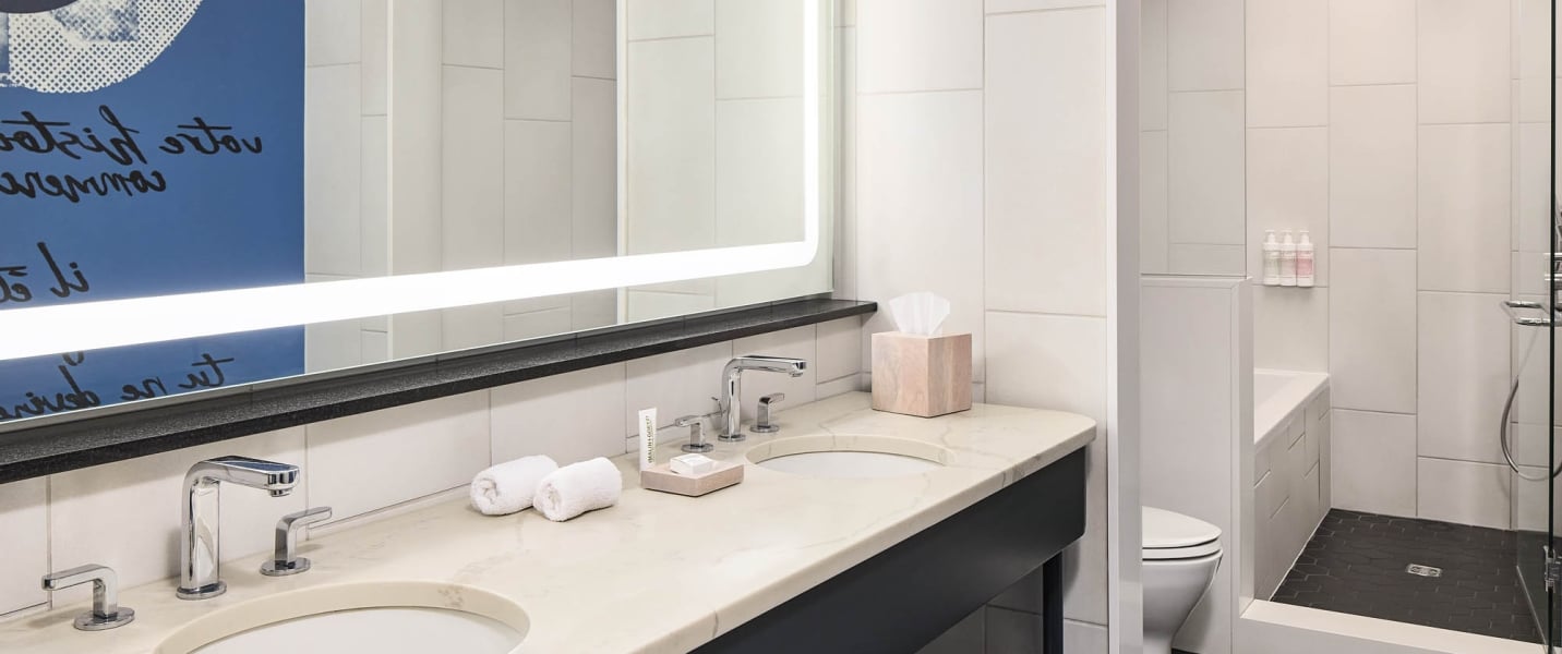 Bathroom vanity features two sinks, chrome faucets, rolled towels, and MAULIN+GOETZ toiletries on a marble counter. A lit mirror reflects blue art. Shower and toilet nearby.