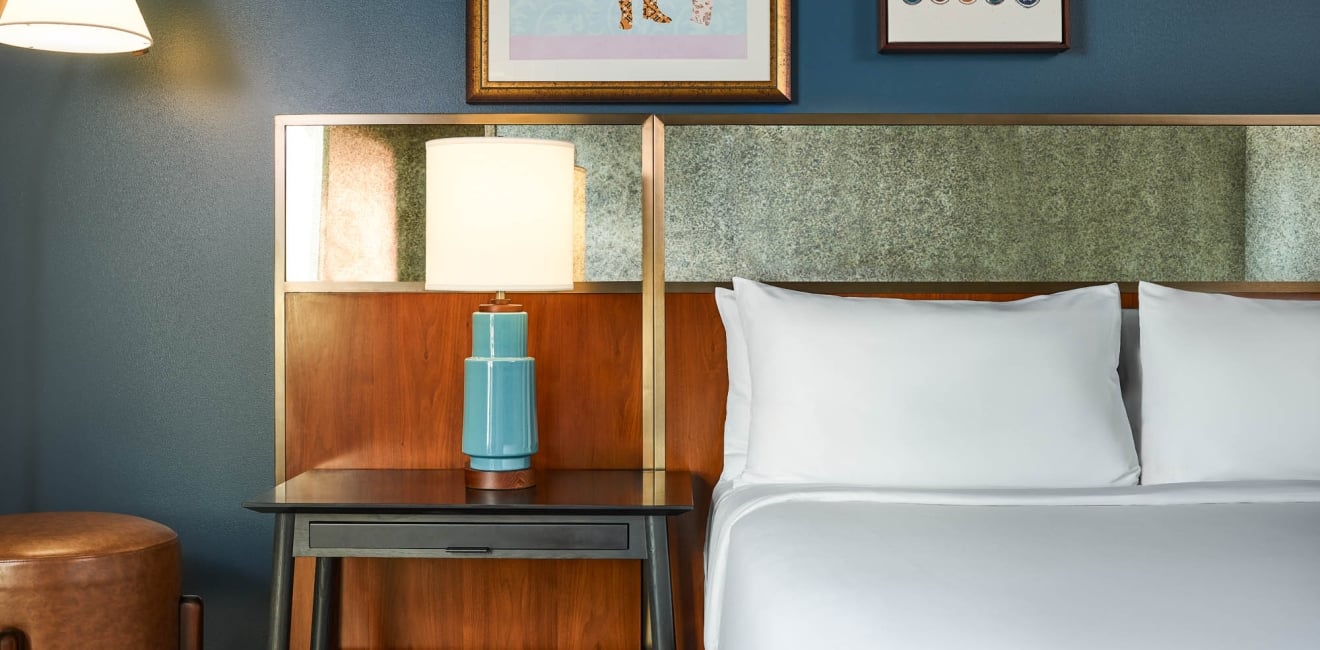 A blue ceramic lamp with a white shade stands on a dark nightstand, beside a bed with white pillows and a textured wood and stone-like headboard.