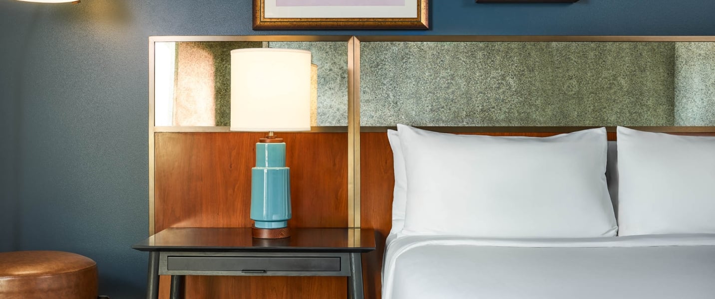 A blue ceramic lamp with a white shade stands on a dark nightstand, beside a bed with white pillows and a textured wood and stone-like headboard.