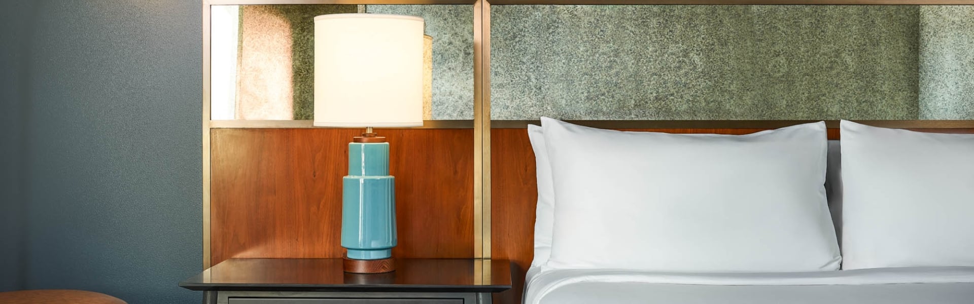 A blue ceramic lamp with a white shade stands on a dark nightstand, beside a bed with white pillows and a textured wood and stone-like headboard.