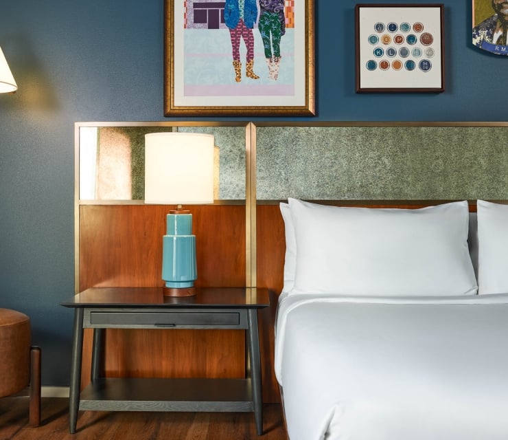 A blue ceramic lamp with a white shade stands on a dark nightstand, beside a bed with white pillows and a textured wood and stone-like headboard.