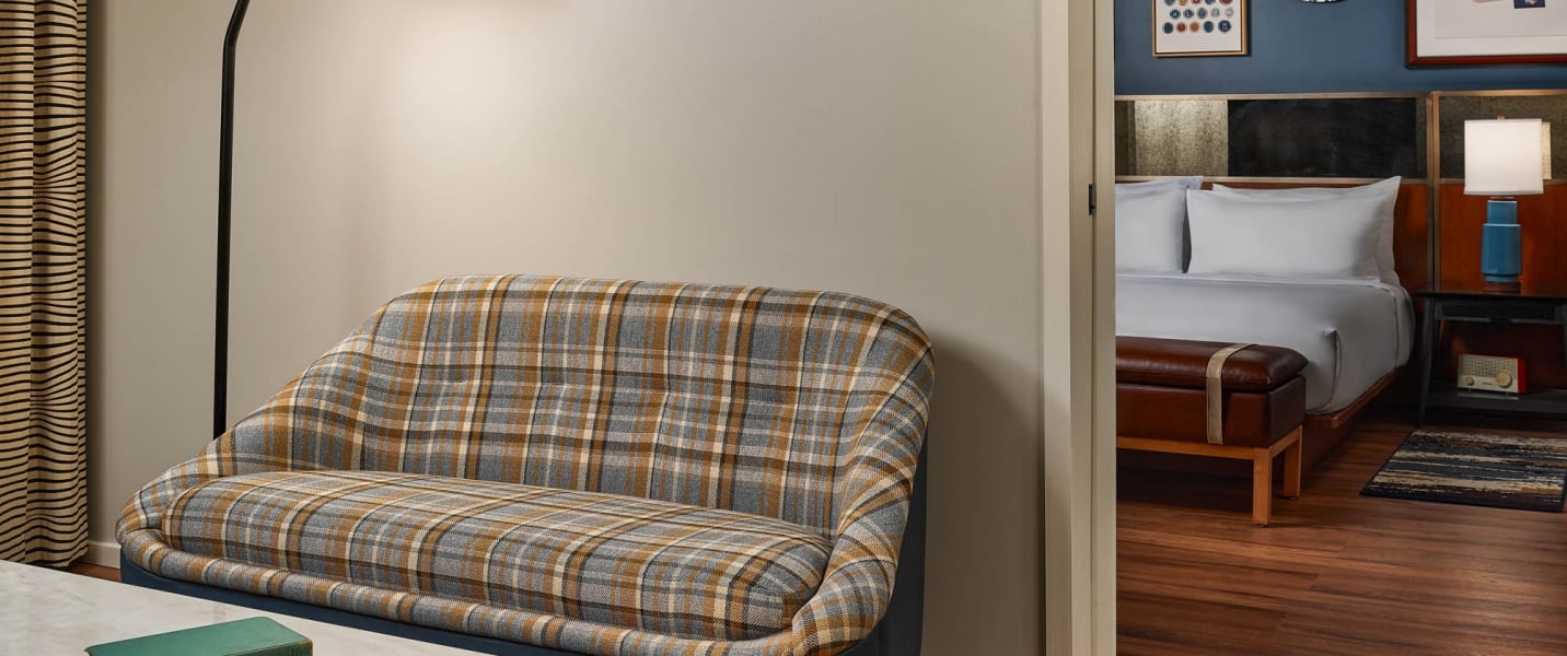 A plaid sofa rests in a room, an open doorway revealing a modern bedroom with a neatly made bed, a leather bench, and a bedside table with a blue lamp.