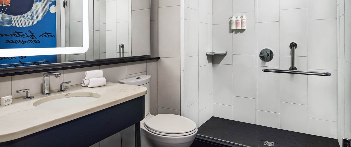 Modern tiled bathroom featuring a sink vanity with towels, a toilet, and a walk-in shower with grab bars. Mirrored text on wall art reads: "vote le destin des hommes".