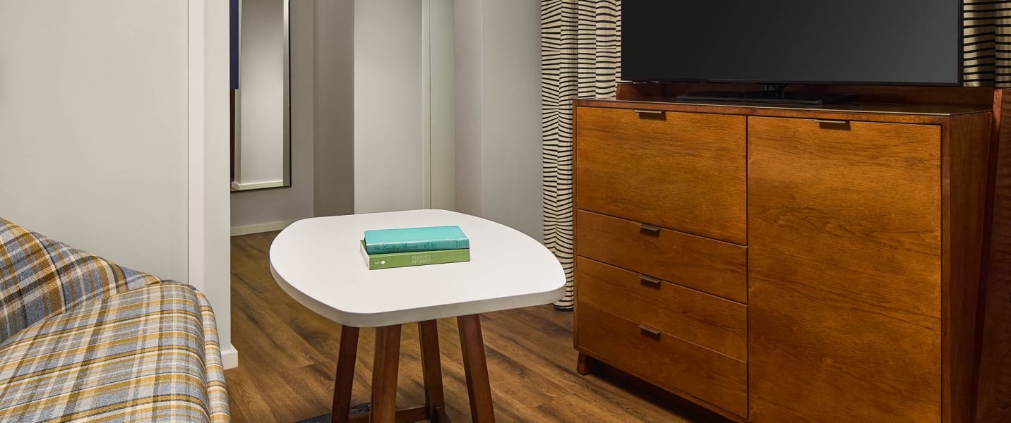 A white coffee table with two "ALBANO AFORO" books sits beside a plaid armchair, facing a wooden cabinet with a TV in a room with light walls and wood floors.