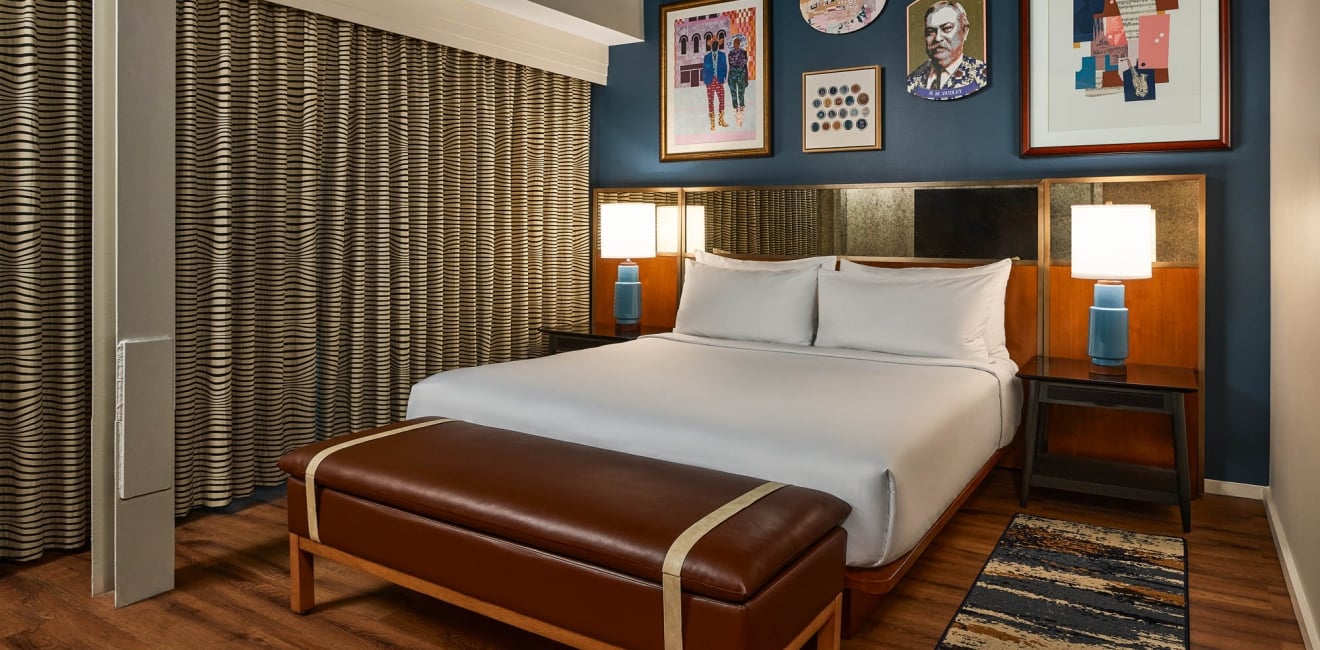 White-sheeted bed is flanked by blue lamps in a modern hotel room with a dark blue wall, framed art, patterned curtains, and a brown leather bench.