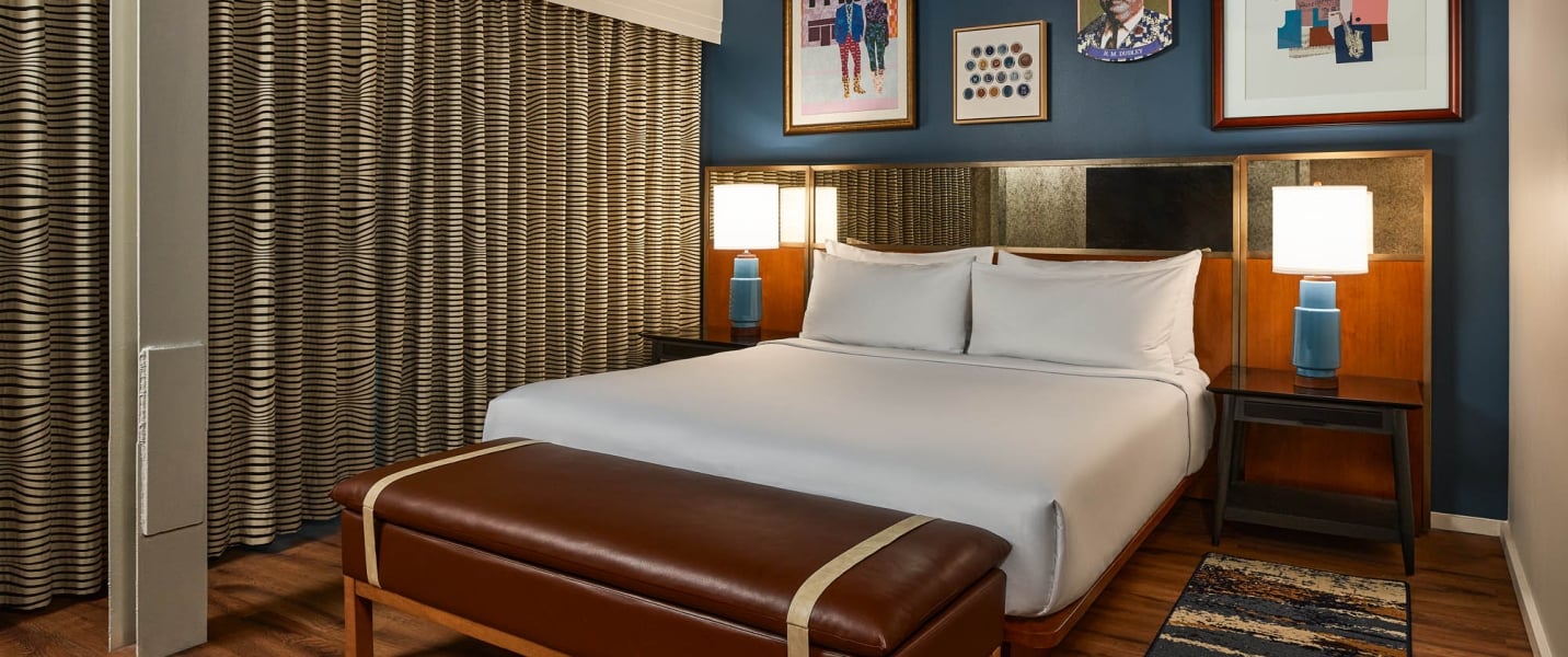 White-sheeted bed is flanked by blue lamps in a modern hotel room with a dark blue wall, framed art, patterned curtains, and a brown leather bench.