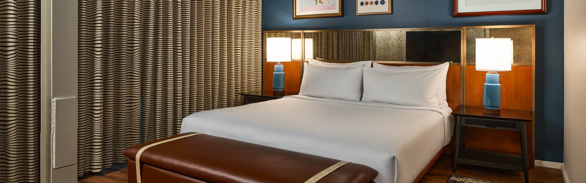 White-sheeted bed is flanked by blue lamps in a modern hotel room with a dark blue wall, framed art, patterned curtains, and a brown leather bench.
