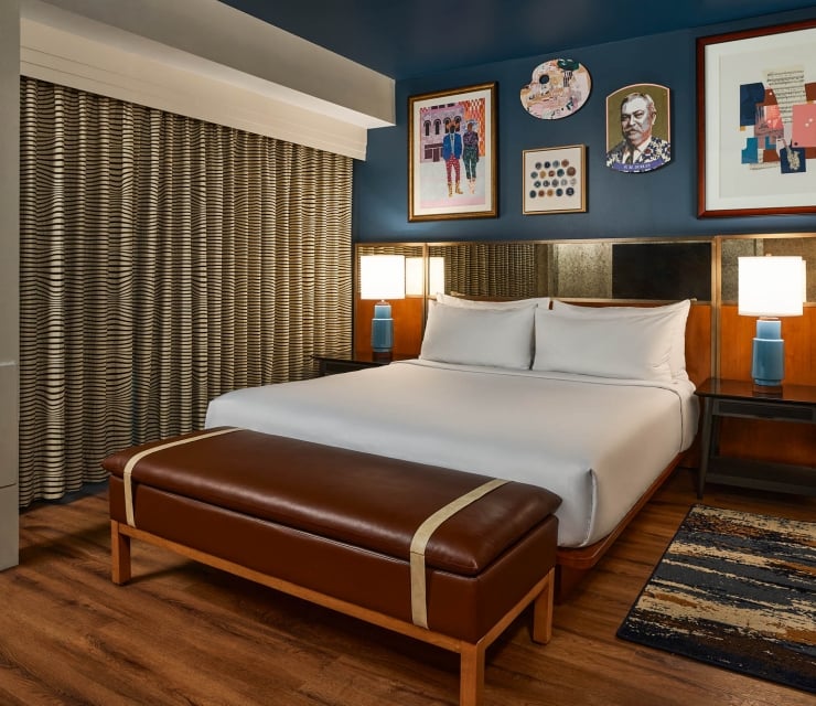 White-sheeted bed is flanked by blue lamps in a modern hotel room with a dark blue wall, framed art, patterned curtains, and a brown leather bench.