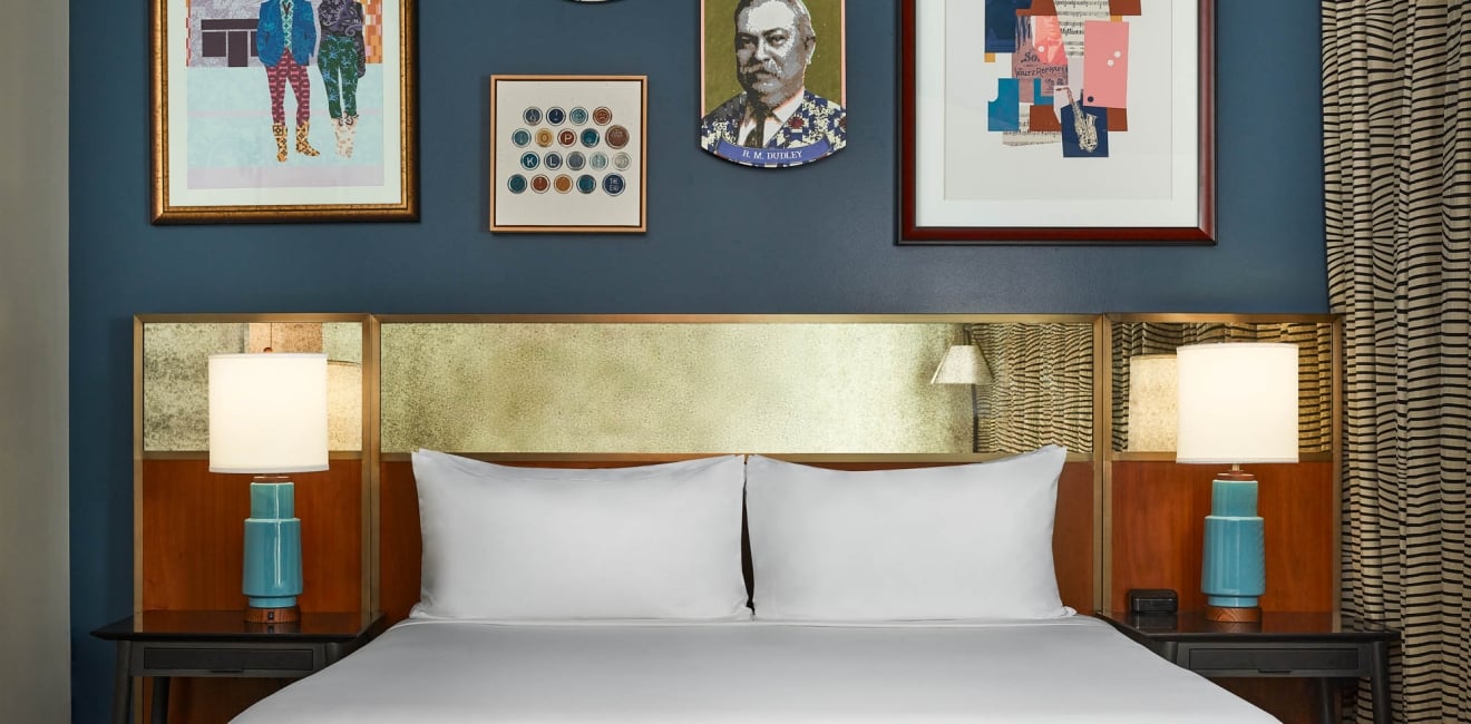 A white-linened bed below a gold headboard, flanked by turquoise lamps. Above, a dark blue wall displays five artworks, including a portrait labeled "H. M. COCKROFT".