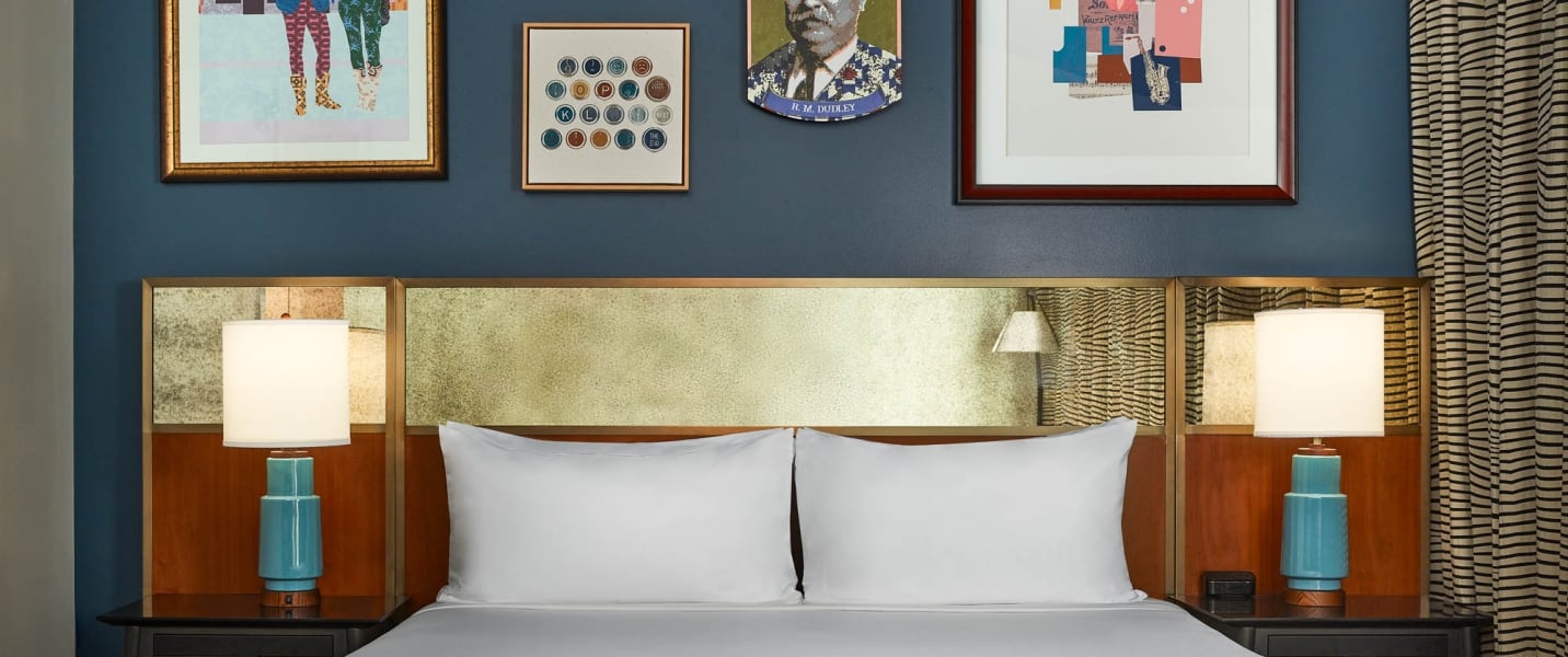 A white-linened bed below a gold headboard, flanked by turquoise lamps. Above, a dark blue wall displays five artworks, including a portrait labeled "H. M. COCKROFT".