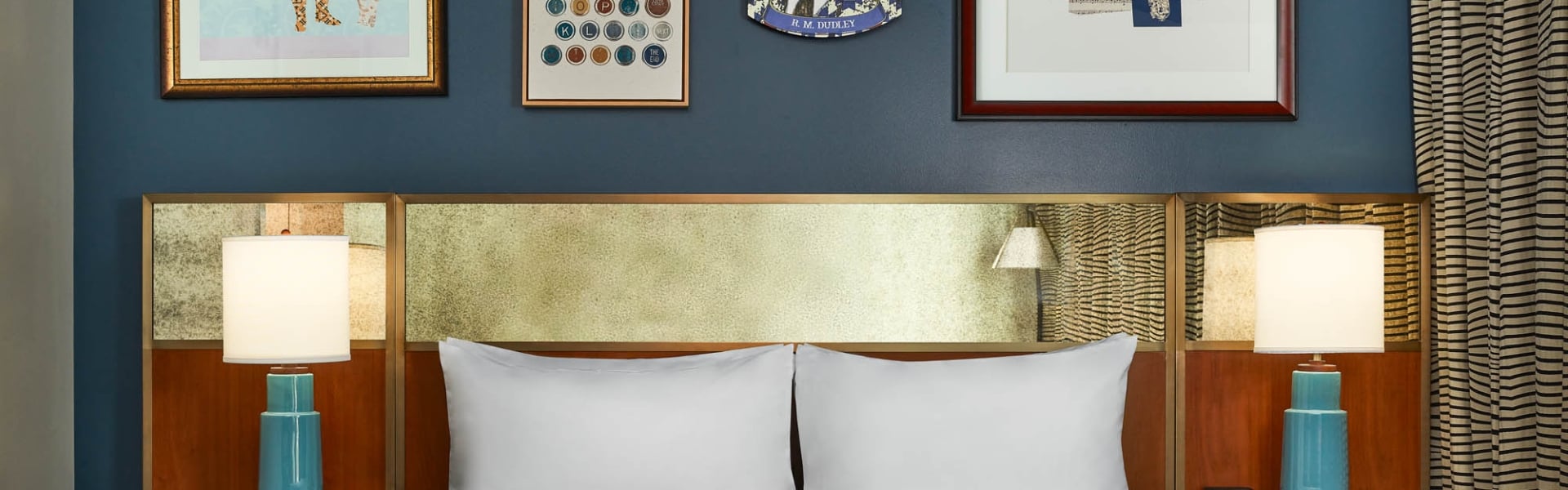 A white-linened bed below a gold headboard, flanked by turquoise lamps. Above, a dark blue wall displays five artworks, including a portrait labeled "H. M. COCKROFT".