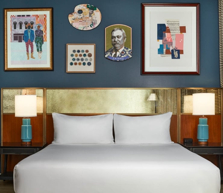 A white-linened bed below a gold headboard, flanked by turquoise lamps. Above, a dark blue wall displays five artworks, including a portrait labeled "H. M. COCKROFT".