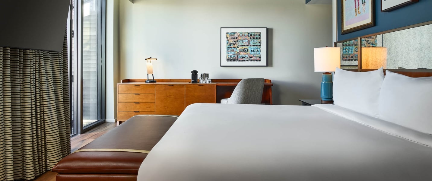 Modern hotel room features a white-linened bed, wooden dresser with coffee maker and desk, plus striped curtains next to a large window.