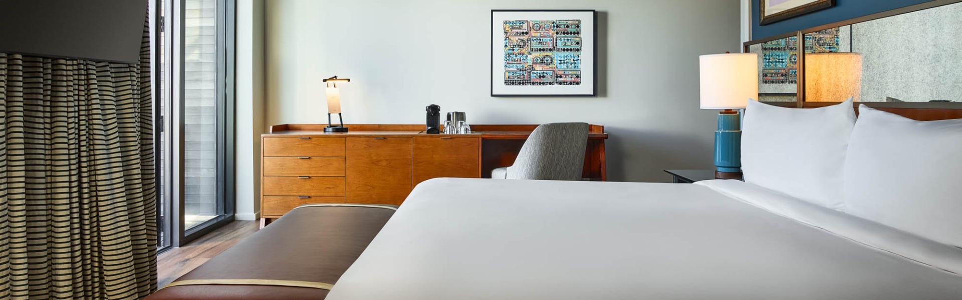 Modern hotel room features a white-linened bed, wooden dresser with coffee maker and desk, plus striped curtains next to a large window.