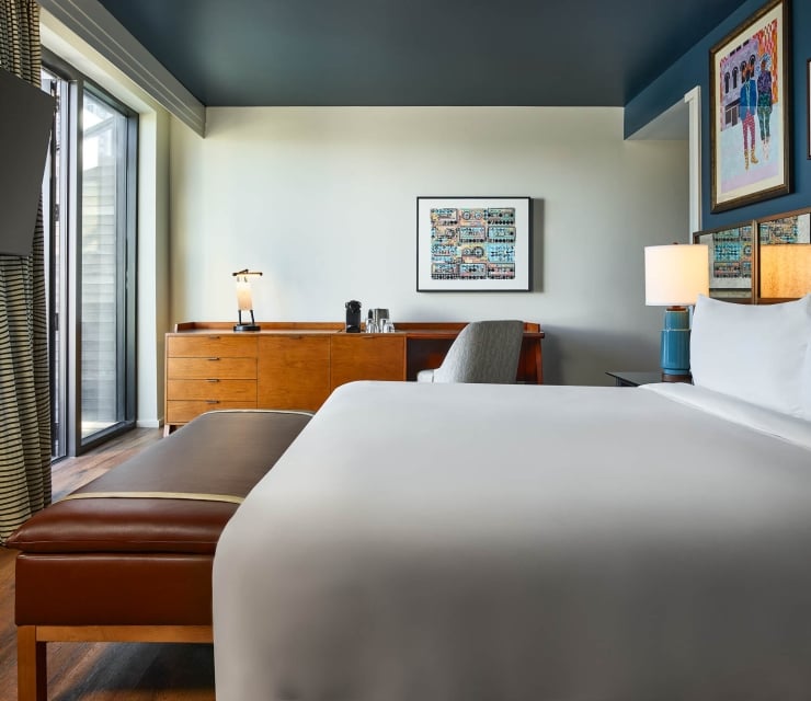 Modern hotel room features a white-linened bed, wooden dresser with coffee maker and desk, plus striped curtains next to a large window.