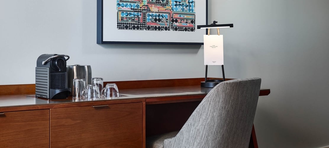 A coffee machine, glassware, and a lamp with "THE UNTITLED HOTEL" sign sit on a wooden desk, beneath abstract art, with a textured gray chair pulled up.