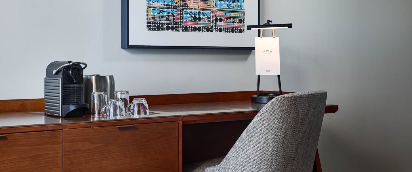 A coffee machine, glassware, and a lamp with "THE UNTITLED HOTEL" sign sit on a wooden desk, beneath abstract art, with a textured gray chair pulled up.