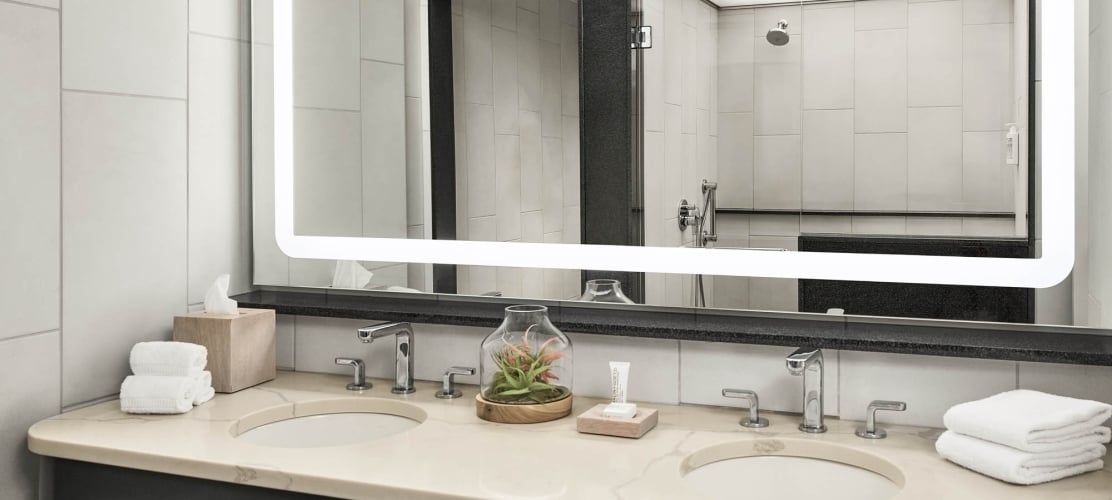 Double vanity and illuminated mirror reflect a tiled shower. Countertop features two sinks, modern faucets, folded white towels, tissue box, a small plant, and a lotion tube.