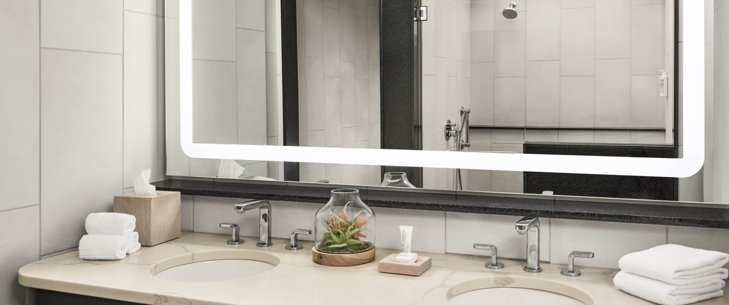 Double vanity and illuminated mirror reflect a tiled shower. Countertop features two sinks, modern faucets, folded white towels, tissue box, a small plant, and a lotion tube.