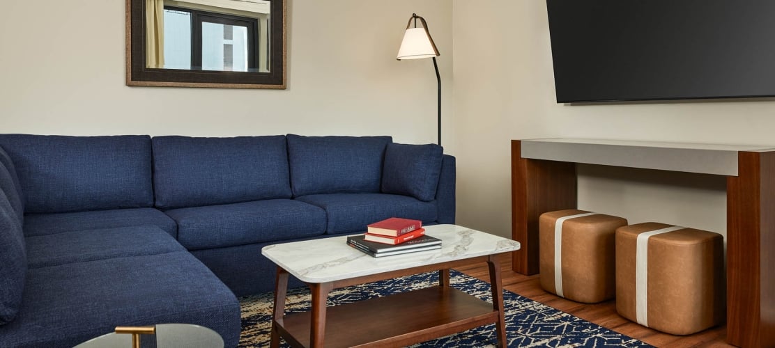 A navy blue sectional sofa furnishes a modern living area. Books sit on a marble coffee table, with ottomans under a console and a TV above, all on a patterned rug.