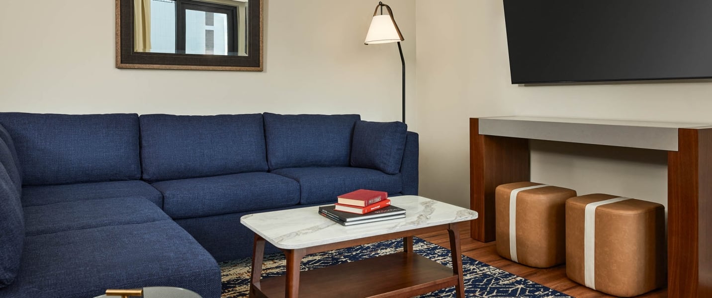 A navy blue sectional sofa furnishes a modern living area. Books sit on a marble coffee table, with ottomans under a console and a TV above, all on a patterned rug.
