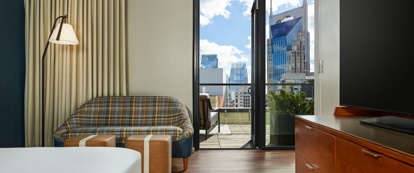 A modern room with a plaid sofa offers a balcony view of a city skyline, including large skyscrapers under a blue sky.