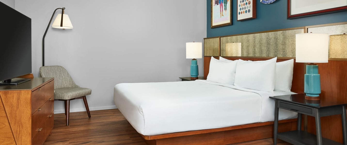 A made bed with white linens is centered in a modern hotel room, flanked by lamps, a TV, and chair, against grey and teal walls with art.