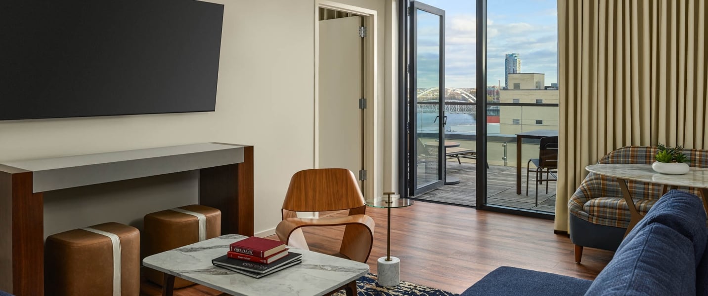 Modern room featuring a TV, coffee table with "FREE TERPIL" book, and various seating. An open door leads to a balcony with city, river, and bridge views.