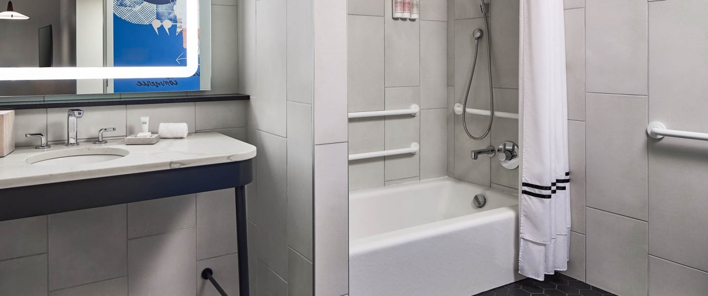 A modern bathroom displays a white marble vanity with an illuminated mirror and a white bathtub-shower combo with grab bars, all set in a light grey tiled room.